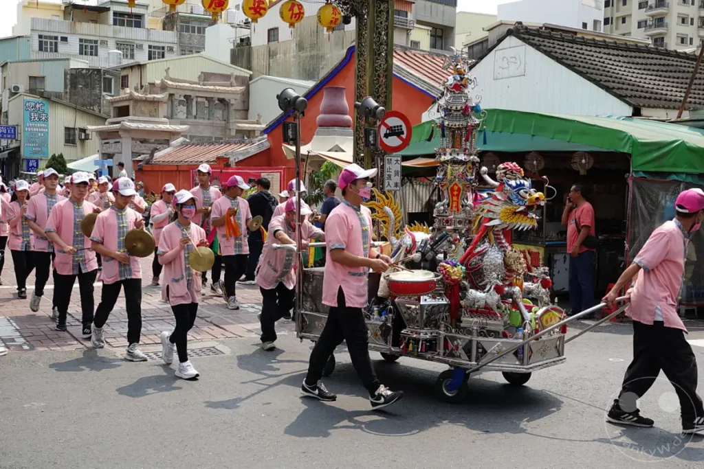 Eine Gruppe von Menschen in pinken Uniformen und Kappen nimmt an einer Prozession in Tainan, Taiwan, teil. Sie spielen Schlaginstrumente und tragen einen reich verzierten Wagen mit einem bunten Drachen darauf, der Teil eines Wind Tempels ist. Die lebendige und feierliche Atmosphäre deutet auf ein kulturelles oder religiöses Fest hin.