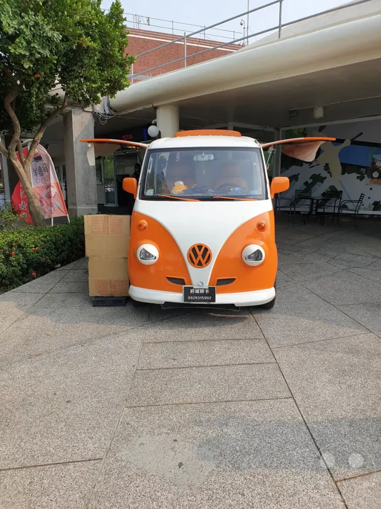 Ein auffälliger, orange-weißer VW-Bus im Retro-Stil steht auf einem Granitpflasterweg. Der Bus ist an einer überdachten Stelle geparkt, mit Sitzgelegenheiten im Hintergrund und einer grünen Baumkulisse auf der linken Seite. Die Szene vermittelt eine entspannte Atmosphäre, möglicherweise an einem Imbissstand oder einer Raststätte in Taiwan.