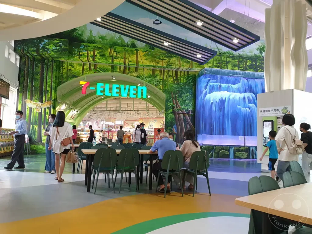 Menschen entspannen in einem modern gestalteten Essbereich einer Autobahnraststätte in Taiwan. Die Umgebung ist von einer üppigen Waldmalerei und einem beeindruckenden Wasserfall dominiert, die eine natürliche Atmosphäre schaffen. Der zentrale 7-Eleven-Markt bietet eine bequeme Einkaufsmöglichkeit inmitten dieser entspannten Kulisse.