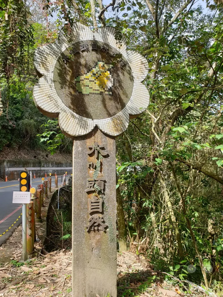 Ein wettergegerbter Gedenkstein mit einer Mosaikskulptur eines Froschkopfes steht aufrecht inmitten üppiger Vegetation. Im Hintergrund ist eine Straße mit Warnleuchten und orangen Kegeln zu sehen, die auf die natürliche Umgebung von Taiwan hinweist. Die Szene strahlt eine ruhige und naturnahe Atmosphäre aus, typisch für die Landschaften rund um den Sun Moon Lake.