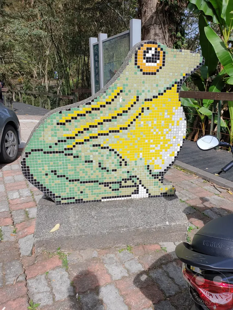 Eine bunte Mosaikskulptur eines Froschkopfes steht auf einem Sockel in einer Parkanlage. Im Hintergrund sind Bäume und Informationstafeln zu sehen, während im Vordergrund ein Roller parkt. Die Sonne wirft Schatten auf den gepflasterten Weg und beleuchtet die Szenerie in Taiwan am Sun Moon Lake.
