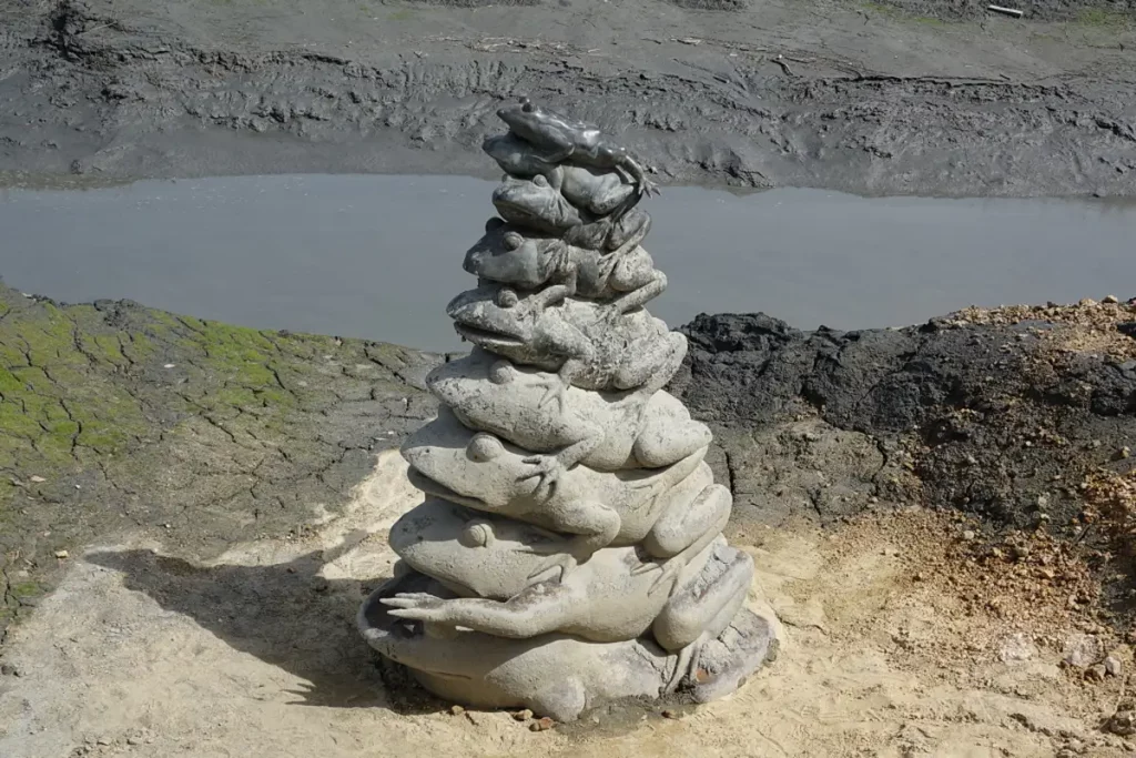 Eine skurrile Steinskulptur aus übereinander gestapelten Fröschen ragt vor der Kulisse eines trüben Gewässers und schlammiger Uferlandschaft in Taiwan auf. Die kunstvolle Anordnung von "Frog Head" und "Frog Sculpture" bildet einen faszinierenden Blickfang am Ufer des "Sun Moon Lake". Die Szene strahlt eine ruhige und leicht bizarre Atmosphäre aus, die von der natürlichen Umgebung eingerahmt wird.