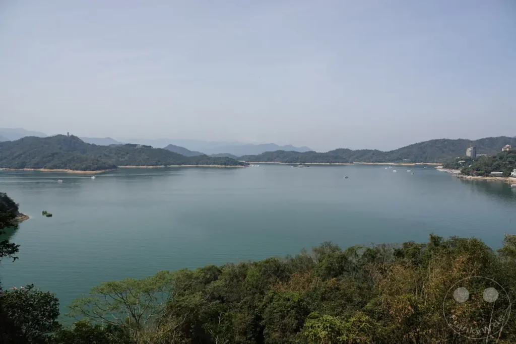 Ein weiter Blick über den malerischen Sun Moon Lake in Taiwan, umgeben von sanften, bewaldeten Bergen. Zahlreiche Boote gleiten auf dem ruhigen, türkisfarbenen Wasser und schaffen eine friedliche Atmosphäre. Die Szene strahlt eine natürliche Schönheit und Gelassenheit aus.