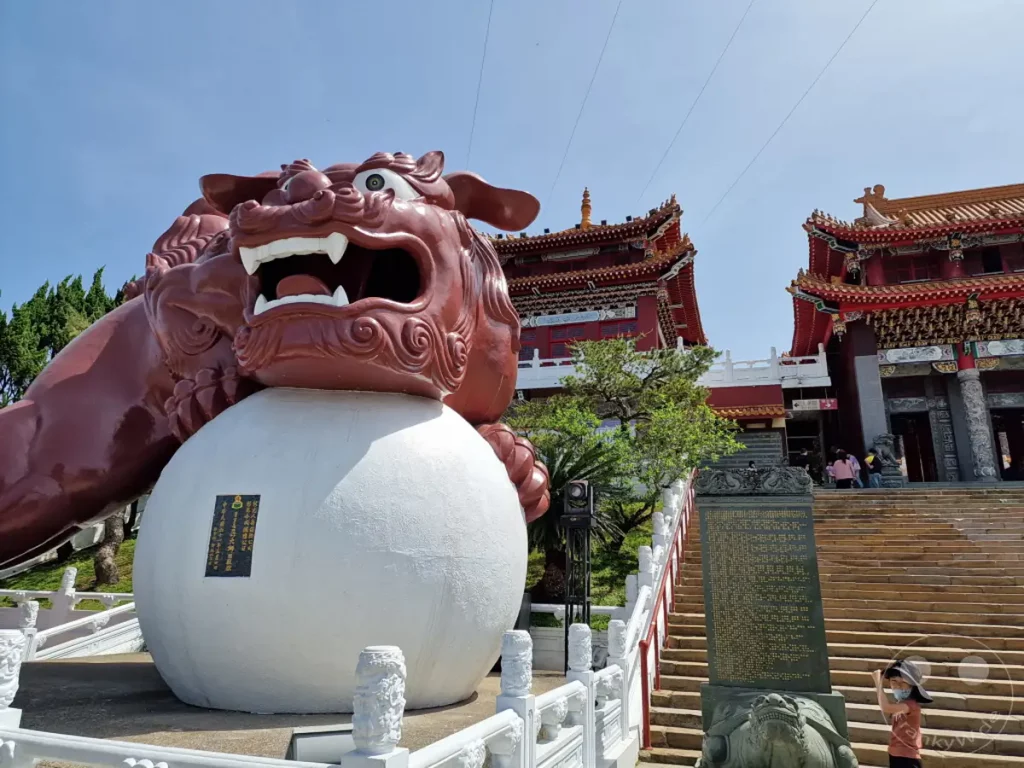 Ein imposanter rotbrauner Löwe steht auf einer großen weißen Kugel vor dem Wenwu Tempel am Sun Moon Lake in Taiwan. Die traditionelle asiatische Architektur des Tempels mit seinen roten Dächern und verzierten Fassaden hebt sich deutlich gegen den blauen Himmel ab. Eine Steintreppe führt zum Haupteingang, während Besucher die kulturelle und spirituelle Atmosphäre dieses malerischen Ortes genießen.