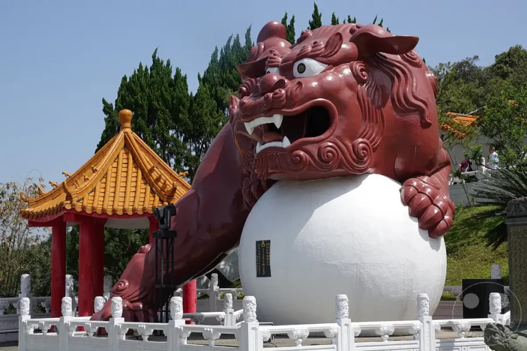 In Taiwan, am malerischen Sun Moon Lake, steht der Wenwu Tempel, dessen Umgebung von eindrucksvollen Skulpturen geschmückt ist. Eine imposante, rotbraune Statue eines Drachenlöwen thront majestätisch über einer weißen Kugel und strahlt eine kraftvolle, mythische Präsenz aus. Hinter der Statue erhebt sich eine traditionelle chinesische Tempelarchitektur mit geschwungenen Dächern und detailreichen Verzierungen, die eine friedliche und zugleich ehrfurchtgebietende Atmosphäre schafft.