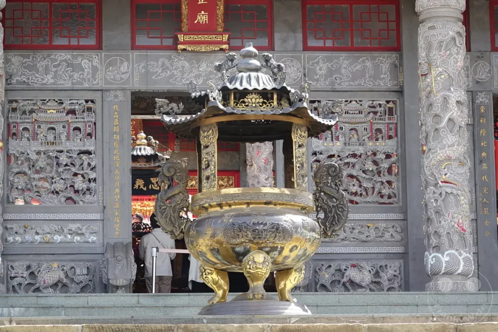 Vor dem Wenwu Tempel am Sun Moon Lake in Taiwan steht ein kunstvoll verziertes, goldenes Räuchergefäß, flankiert von detaillierten Drachenskulpturen. Die reich verzierte Fassade des Tempels mit filigranen Holzschnitzereien und roten Fenstern bildet einen beeindruckenden Hintergrund. Die Szene strahlt eine ruhige und ehrwürdige Atmosphäre aus, die die kulturelle und spirituelle Bedeutung des Ortes unterstreicht.