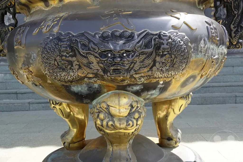 Ein imposanter Bronzekessel mit detailreichen Drachenreliefs und chinesischen Schriftzeichen steht vor einer Treppenanlage im Wenwu Tempel am Sun Moon Lake in Taiwan. Die Patina des Kessels deutet auf sein Alter und seine Wichtigkeit hin, während die goldfarbenen Löwenfüße und die scharfen Drachenaugen eine majestätische und ehrwürdige Atmosphäre schaffen.