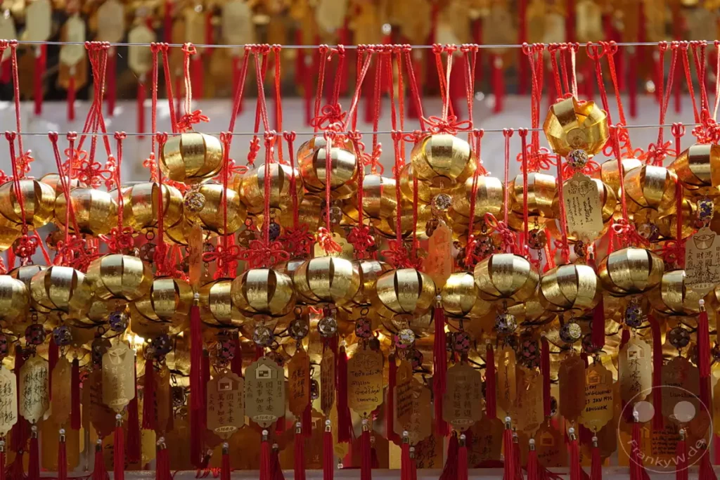 Goldfarbene Glücksbringer und Gebetsanhänger hängen in Reihen an roten Schnüren vor dem Wenwu Tempel am Sun Moon Lake in Taiwan. Die zahlreichen kleinen, glänzenden Objekte sind mit Schriftzeichen verziert und strahlen eine festliche und spirituelle Atmosphäre aus. Sie vermitteln ein Gefühl von Hoffnung und Segen, das typisch für religiöse Stätten in dieser Region ist.