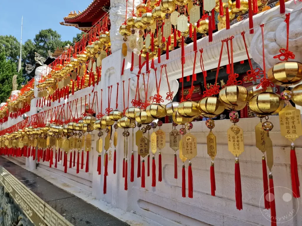Zahlreiche goldene Glücksbringer mit roten Quasten hängen an einer weißen Mauer des Wenwu Tempels am Sun Moon Lake in Taiwan. Die kunstvollen Verzierungen und Anhänger schaffen eine festliche und spirituelle Atmosphäre. Die leuchtenden Farben und die schiere Menge der Glücksbringer erzeugen ein beeindruckendes visuelles Erlebnis vor der Kulisse des Tempels und der Natur.