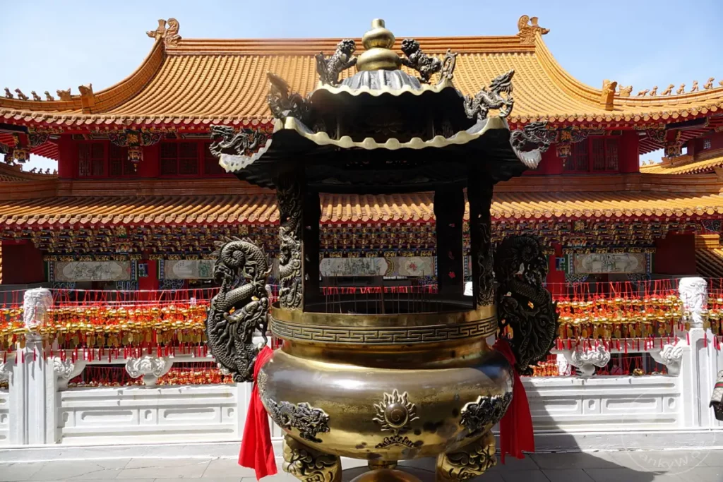 Ein imposanter bronzener Räucherofen mit detaillierten Drachenverzierungen steht im Vordergrund vor der prächtigen Architektur des Wenwu Tempels am Sun Moon Lake in Taiwan. Zahlreiche goldene Amulette mit roten Bändern hängen an einer Balustrade und verleihen der Szene eine festliche und spirituelle Atmosphäre. Die roten und gelben Farben des Tempels und die klaren Linien der Dächer spiegeln die traditionelle chinesische Bauweise wider.