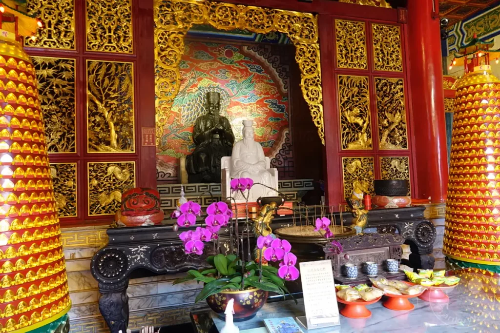 Im Wenwu Tempel am Sun Moon Lake in Taiwan stehen prächtige Statuen von Buddha und anderen Figuren auf kunstvoll gestalteten Altären, umgeben von goldenen Verzierungen und roten Säulen. Helle Orchideenblüten und Räucherstäbchen schaffen eine friedliche und spirituelle Atmosphäre im Inneren des Tempels. Die kunstvollen Schnitzereien und die leuchtenden Farben der Dekorationen unterstreichen die religiöse Bedeutung und die traditionelle Architektur.
