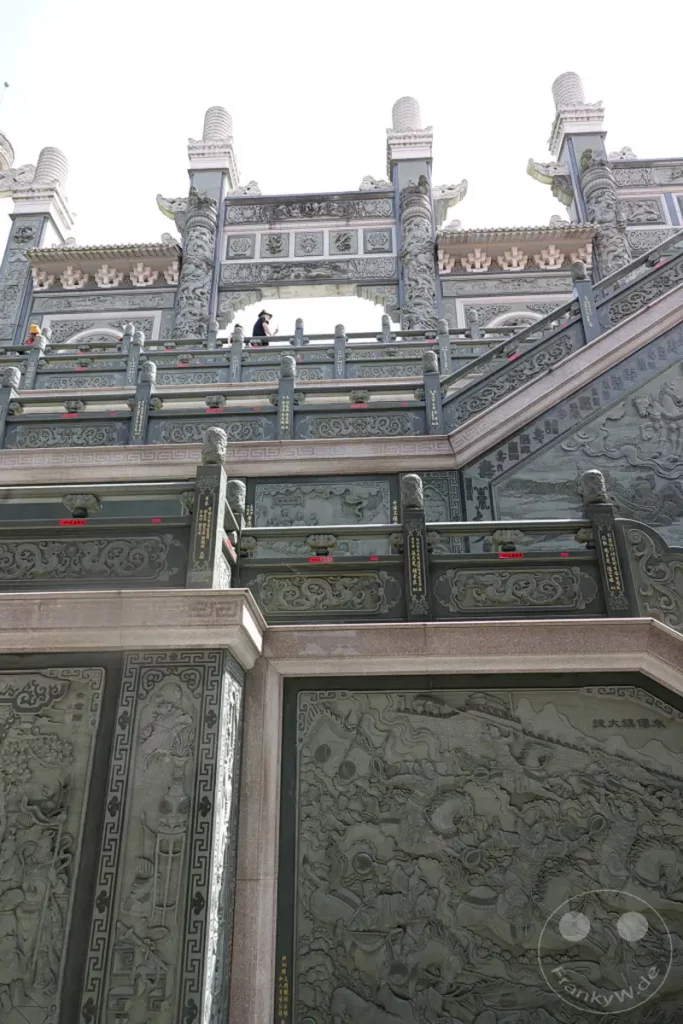 Ein weitläufiger Blick auf die imposante Tempelanlage des Wenwu Tempels am Sun Moon Lake in Taiwan, dessen Fassaden mit kunstvollen Steinmetzarbeiten verziert sind. Die Architektur im traditionell asiatischen Stil erzeugt eine ehrwürdige und kulturell reiche Atmosphäre. Auf einer oberen Ebene ist eine Person zu sehen, die die Aussicht genießt.