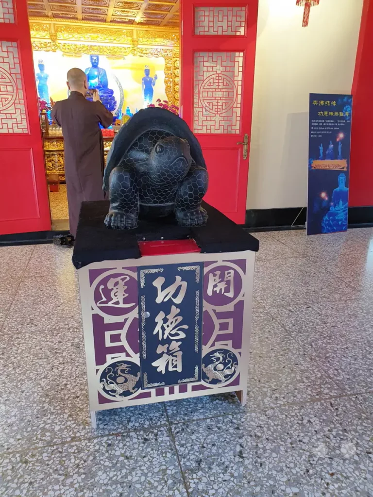 Im Wenwu Temple am Sun Moon Lake in Taiwan steht eine große Schildkrötenstatue vor einem Altar mit Buddha-Statuen, während ein Mönch im Hintergrund eine Opfergabe darbringt. Die Szene strahlt eine ruhige und spirituelle Atmosphäre aus, die von den leuchtenden Farben und der detailreichen Gestaltung der Artefakte unterstrichen wird. Ein blaues Schild mit chinesischen Schriftzeichen und buddhistischen Motiven ergänzt die mystische Stimmung dieses religiösen Ortes.