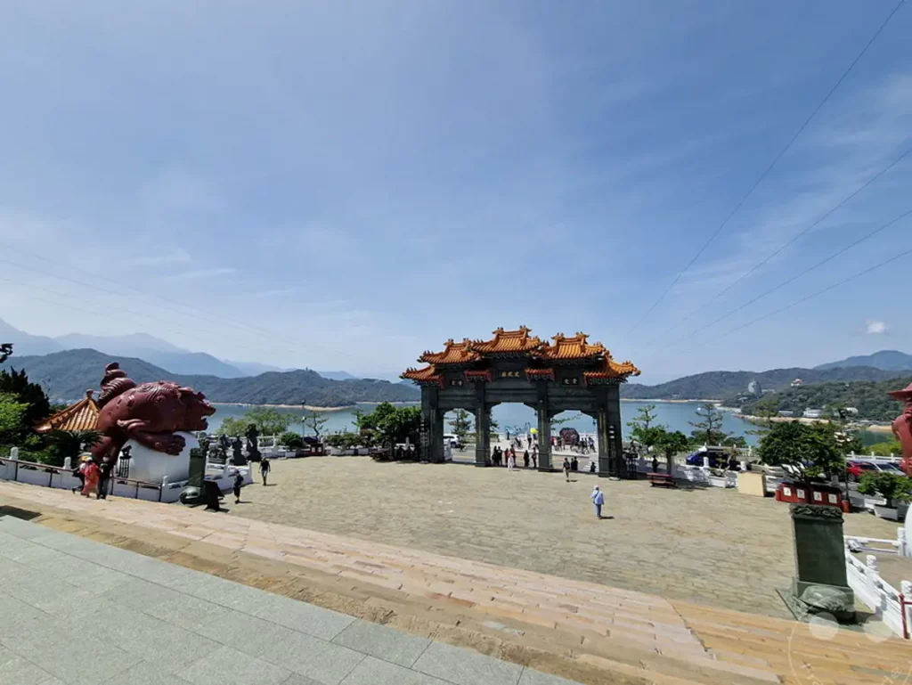 Das Bild vermittelt eine friedliche Atmosphäre am Ufer des Sun Moon Lake in Taiwan. Die beeindruckende Architektur des Wenwu Tempels mit seinen leuchtend orangefarbenen Dächern dominiert die Szenerie, während im Hintergrund sanfte Berge aufragen. Touristen genießen die weite Aussicht auf den See und die umliegende Natur unter einem strahlend blauen Himmel.