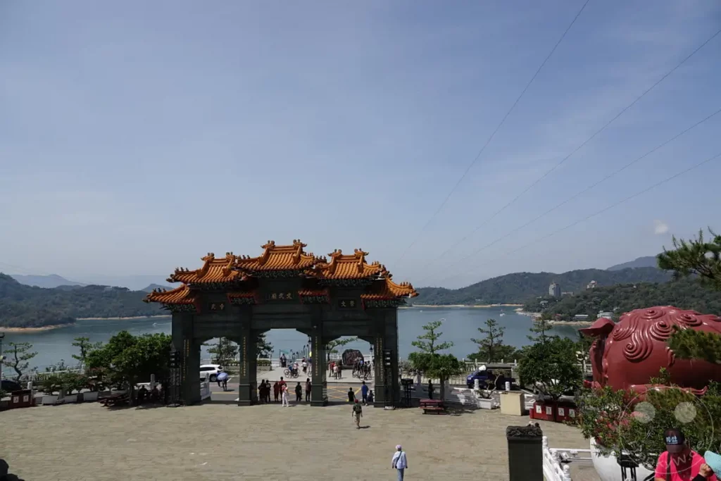 Die imposante Toranlage des Wenwu Tempels am Sun Moon Lake in Taiwan thront über einem weitläufigen Platz mit Blick auf den ruhigen See und die umliegenden Berge. Zahlreiche Besucher schlendern durch den Bereich und genießen die sonnige Atmosphäre. Ein großer, roter Tempel Löwe wacht über den Eingang, während im Hintergrund die malerische Landschaft des Sun Moon Lake und bewaldete Hügel zu sehen sind.
