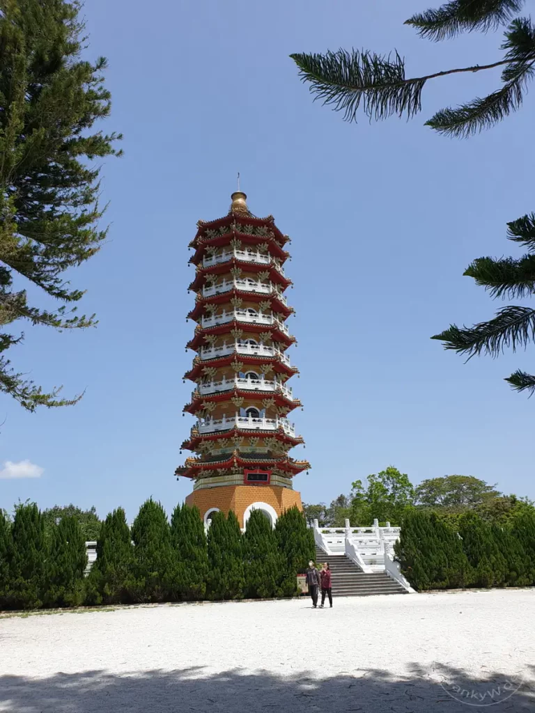 Die Ci'en Pagoda erhebt sich majestätisch in den blauen Himmel über dem Sun Moon Lake in Taiwan. Umgeben von immergrünen Bäumen und mit einer weißen Steintreppe, die zum Eingang führt, strahlt die traditionelle chinesische Architektur Ruhe und Erhabenheit aus. Zwei Besucher spazieren auf dem Kiesweg und bewundern die imposante Sehenswürdigkeit bei strahlendem Sonnenschein.