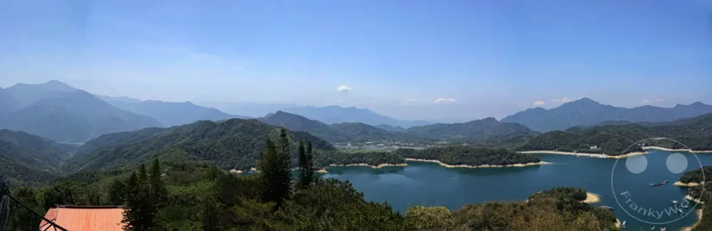 Ein weitläufiges Panorama mit dem tiefblauen Sun Moon Lake im Vordergrund, umgeben von üppig grünen Bergen in Taiwan. Die klare Luft und der strahlend blaue Himmel schaffen eine friedliche und majestätische Atmosphäre, die zum Verweilen einlädt. Im Hintergrund erheben sich sanfte Hügelketten und ferne Berge, die die natürliche Schönheit der Region unterstreichen.