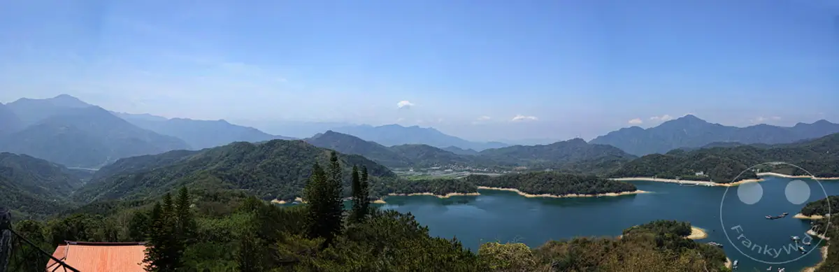 Ein weitläufiges Panorama mit dem tiefblauen Sun Moon Lake im Vordergrund, umgeben von üppig grünen Bergen in Taiwan. Die klare Luft und der strahlend blaue Himmel schaffen eine friedliche und majestätische Atmosphäre, die zum Verweilen einlädt. Im Hintergrund erheben sich sanfte Hügelketten und ferne Berge, die die natürliche Schönheit der Region unterstreichen.