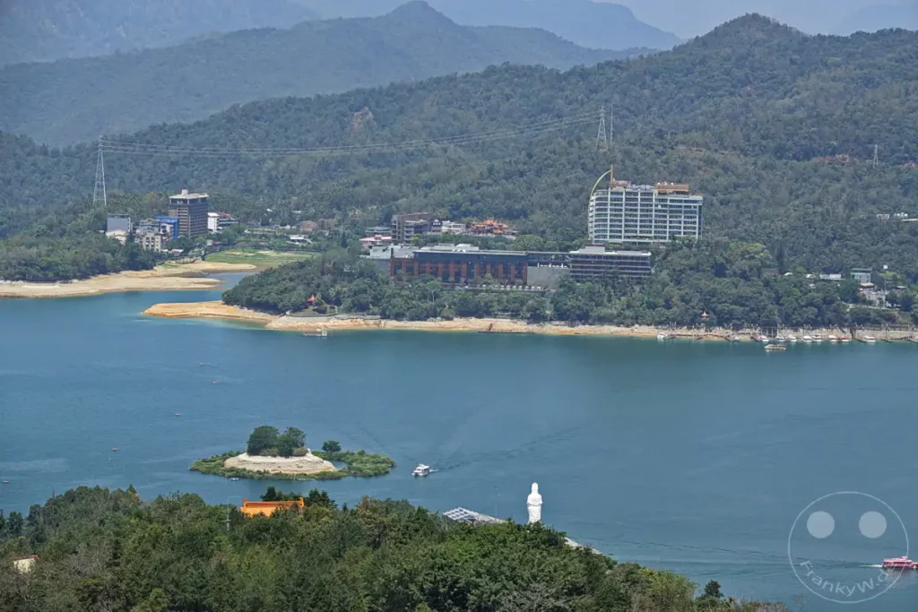 Das Bild fängt eine weite Ansicht des Sun Moon Lake in Taiwan ein, mit grünen Bergen im Hintergrund und verschiedenen Gebäuden am Ufer. Ein kleiner Inselberg mit Bäumen und eine weiße Buddha-Statue dominieren die vordere Wasserfläche, während Boote auf dem ruhigen See unterwegs sind. Die Szene vermittelt eine friedliche und malerische Atmosphäre der Natur und Architektur.