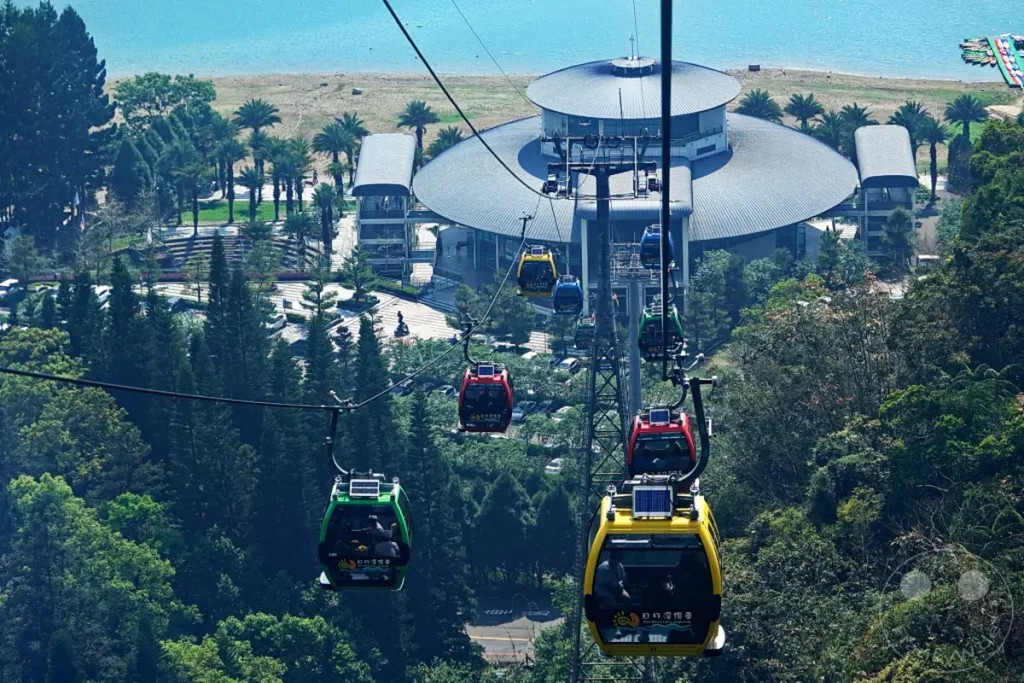 Mehrere bunte Gondeln der Sun Moon Lake Ropeway schweben über einer üppigen Berglandschaft in Taiwan. Im Hintergrund sind die namensgebende Wasserfläche des Sun Moon Lake und die moderne Riyuetanlan Ropeway Station zu erkennen. Die Szene vermittelt ein Gefühl von Erhabenheit und Abenteuer inmitten der natürlichen Schönheit.