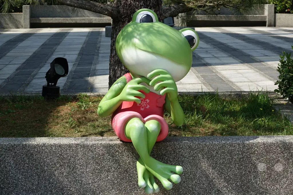 Eine lebensecht wirkende, grüne Frosch-Skulptur sitzt entspannt auf einer steinernen Mauer in der Nähe der Riyuetanlan Seilbahnstation am Sun Moon Lake in Taiwan. Die Skulptur trägt ein pinkfarbenes Kleid mit Blumenmuster und die Beine sind lässig übereinander geschlagen, was eine friedliche und verspielte Atmosphäre am malerischen See erzeugt. Sie ist umgeben von sattem Gras und spendet Schatten spendenden Baum, während im Hintergrund ein gepflasterter Weg und weitere Bänke zu sehen sind.