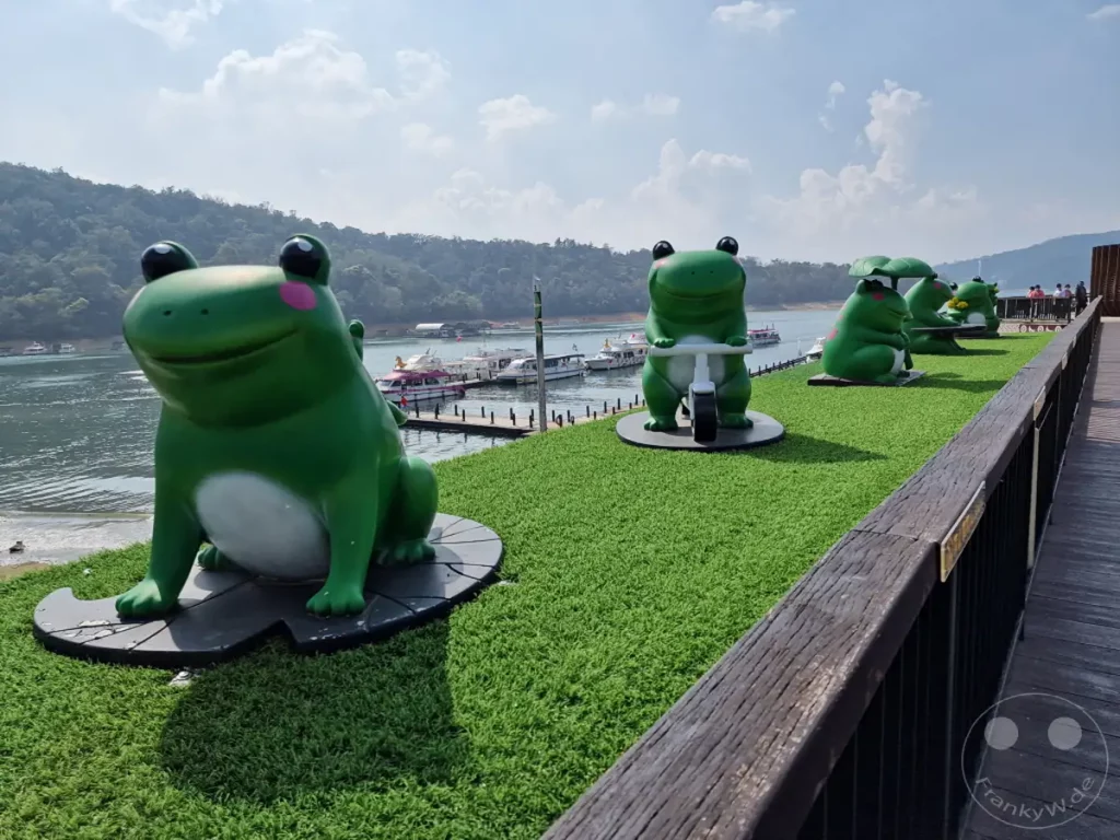 In der Nähe des Yidashao Wharf am Sun Moon Lake in Taiwan stehen mehrere grüne Froschfiguren auf einer künstlichen Grasfläche. Die fröhlich gestalteten Skulpturen blicken auf den See mit Booten und den bewaldeten Hügeln im Hintergrund. Die Szene vermittelt eine entspannte und touristische Urlaubsatmosphäre.
