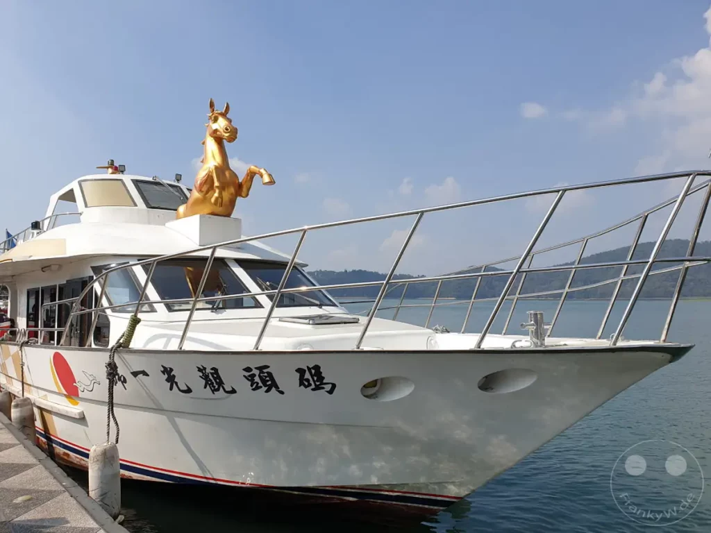 Ein weißes Boot mit einer goldenen Pferdeskulptur auf dem Dach liegt am Ufer des Sun Moon Lake in Taiwan. Die Spiegelung des Bootes und die ruhige See schaffen eine friedliche Atmosphäre. Im Hintergrund erheben sich sanfte Hügel, die die malerische Landschaft des Yidashao Wharf unterstreichen.