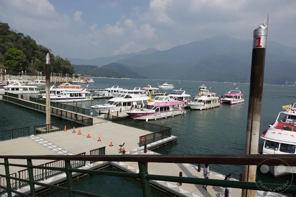 Am Shuishe Wharf am Sun Moon Lake in Taiwan reihen sich zahlreiche Ausflugsboote aneinander, bereit für ihre Fahrten über den malerischen See. Im Hintergrund erheben sich sanfte, bewaldete Berge vor einem leicht bewölkten Himmel, während das ruhige Wasser des Sees die Szenerie widerspiegelt. Eine friedliche und geschäftige Atmosphäre herrscht an diesem beliebten Touristenziel.
