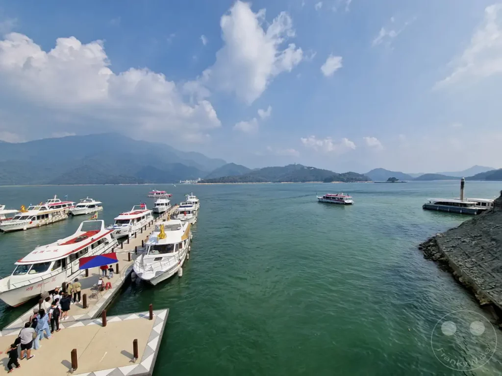 Zahlreiche weiße Ausflugsboote liegen am Shuishe Wharf im Sun Moon Lake in Taiwan an einem sonnigen Tag vor Anker. Im Hintergrund erheben sich sanfte, grüne Berge unter einem strahlend blauen Himmel mit vereinzelten Wolken. Das türkisfarbene Wasser des Sees und die belebte Anlegestelle erzeugen eine lebendige und friedliche Urlaubsatmosphäre.