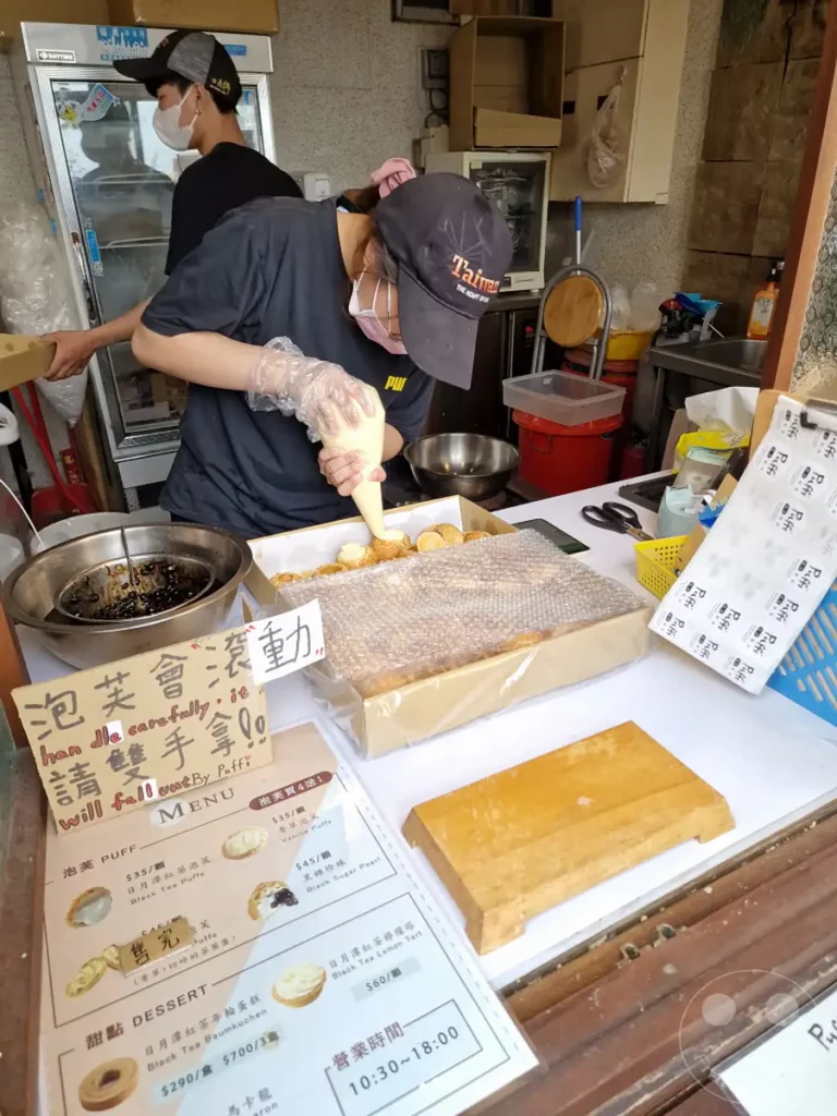 In Shuishe, Taiwan, werden frisch zubereitete Gebäckkreationen von "Puffi" sorgfältig verpackt. Eine Mitarbeiterin füllt mit einer Spritztülle süße Cremes in die kleinen, runden Teiglinge, während im Hintergrund eine Auswahl an feinen Torten und anderen Leckereien zum Verkauf ausliegt. Die Szene strahlt die sorgfältige Handwerkskunst und die köstliche Vielfalt der Bäckerei aus.