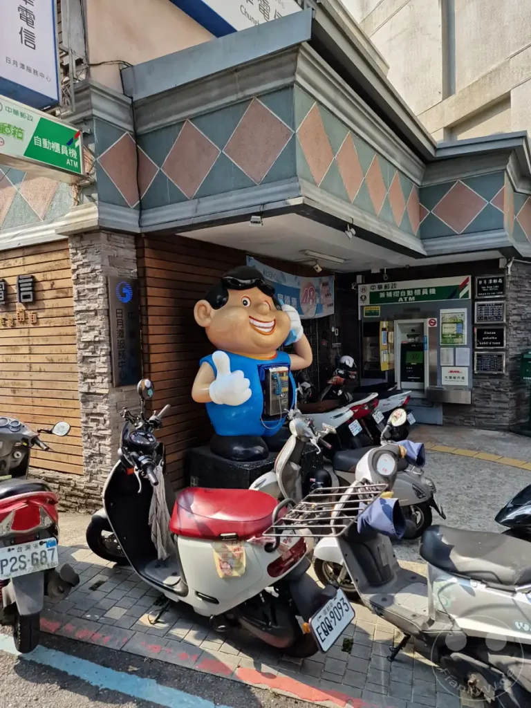 In Shuishe, einem Ort am malerischen Sun Moon Lake in Taiwan, steht ein bunter "Telefonmann" als Werbefigur vor einem Postamt. Mehrere Roller sind ordentlich vor dem Gebäude geparkt, was auf das tägliche Leben und die Fortbewegung in dieser touristischen Gegend hinweist. Die Szene strahlt eine friedliche und alltägliche Atmosphäre aus, beleuchtet von hellem Sonnenschein.