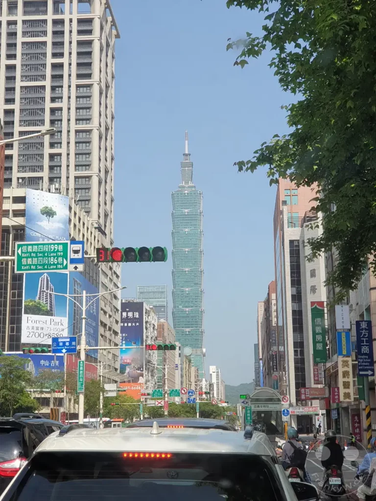 In Taipeh, Taiwan, ragt der ikonische Wolkenkratzer Taipei 101 über die belebte Straßenschlucht empor, während der Verkehr stockt. Die Szene fängt die urbane Dichte und das geschäftige Treiben einer modernen asiatischen Metropole ein. Ein klarer Himmel bildet einen ruhigen Kontrast zur dynamischen städtischen Umgebung.
