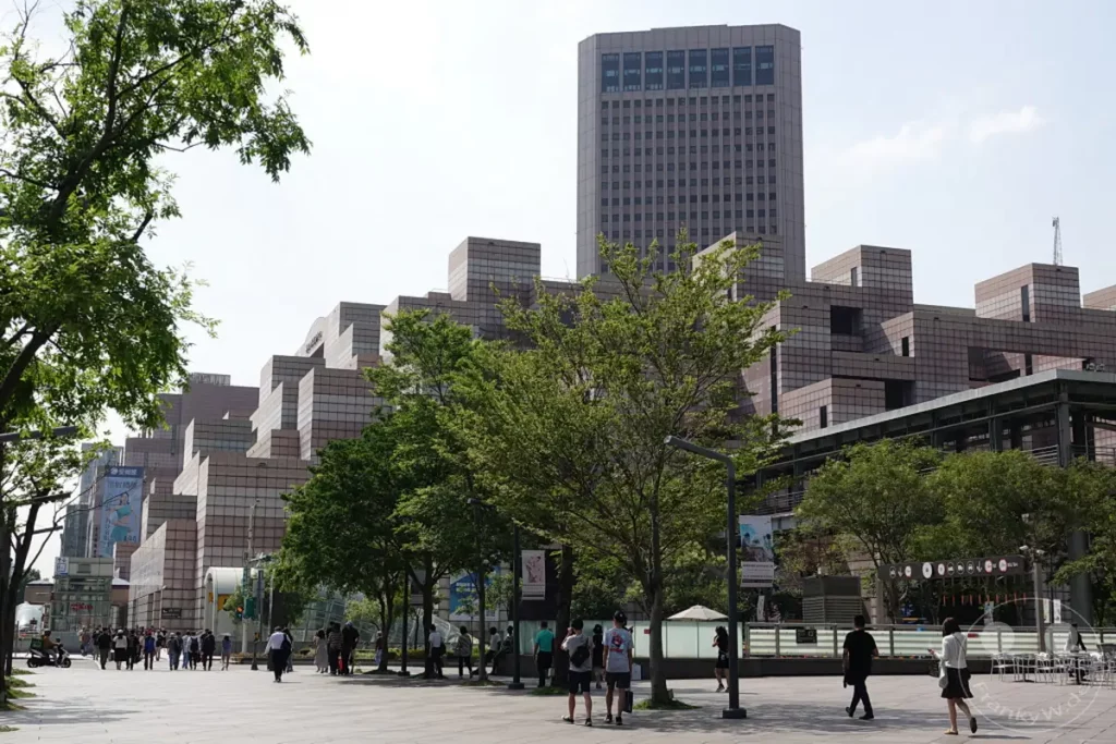Ein weitläufiger Platz in Taipeh, Taiwan, mit modernen Gebäuden und dem imposanten Taipei 101 im Hintergrund. Menschen flanieren auf dem gepflasterten Weg, umgeben von Bäumen und städtischer Architektur. Die Szene vermittelt eine lebendige Atmosphäre an einem sonnigen Tag.