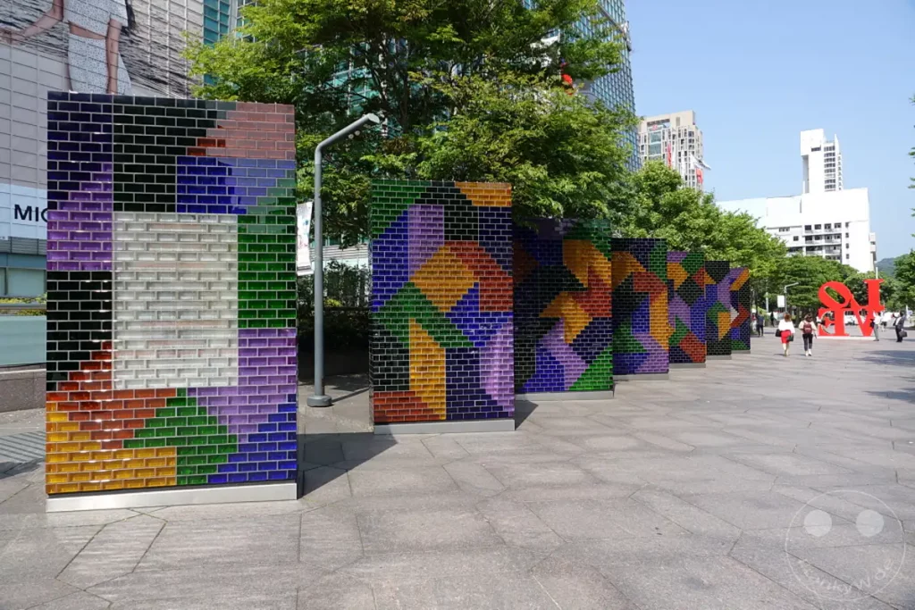 Die leuchtenden, geometrischen Skulpturen aus bunten Glasziegeln säumen einen modernen Platz in Taipeh. Im Hintergrund ragen die charakteristischen Gebäude der Skyline von Taipeh, einschließlich des Taipei 101, in den klaren Himmel. Die Szene vermittelt eine lebendige und urbane Atmosphäre, die von Kunst und Architektur geprägt ist.