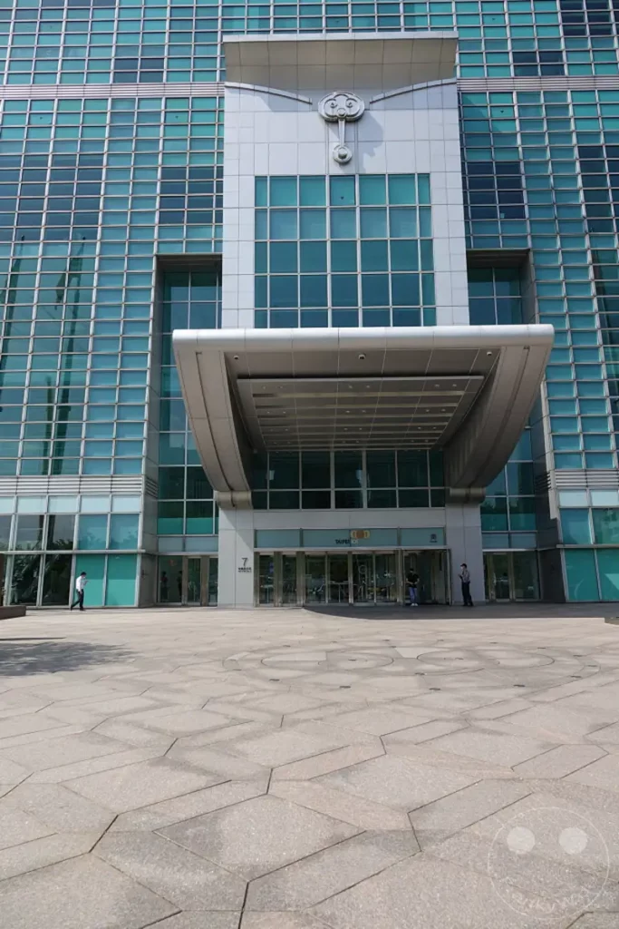 Der imposante Eingangsbereich des Taipei 101 in Taipeh, Taiwan, wird von seiner modernen Glasfassade und einem charakteristischen Vordach dominiert. Die Gestaltung des Platzes mit seinen geometrischen Steinplatten und die klare Sonneneinstrahlung vermitteln eine Atmosphäre von urbaner Größe und Ordnung. Mehrere Personen bewegen sich auf dem weitläufigen Vorplatz, was dem architektonischen Ensemble Leben einhaucht.