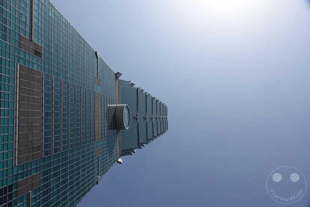 Der Taipei 101, ein ikonischer Wolkenkratzer in Taipeh, Taiwan, ragt empor gegen einen strahlend blauen Himmel. Seine moderne Glasfassade reflektiert das klare Tageslicht und betont die beeindruckende architektonische Leistung. Die Perspektive von unten vermittelt ein Gefühl von Größe und Ehrfurcht vor diesem architektonischen Wahrzeichen.