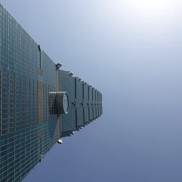 Der Taipei 101, ein ikonischer Wolkenkratzer in Taipeh, Taiwan, ragt empor gegen einen strahlend blauen Himmel. Seine moderne Glasfassade reflektiert das klare Tageslicht und betont die beeindruckende architektonische Leistung. Die Perspektive von unten vermittelt ein Gefühl von Größe und Ehrfurcht vor diesem architektonischen Wahrzeichen.