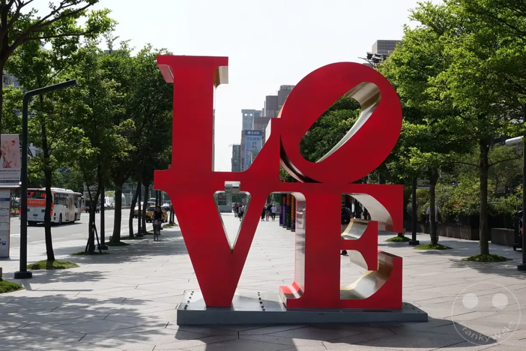Die ikonische rote "LOVE" Skulptur steht prominent auf einem belebten Gehweg in Taipeh, Taiwan. Im Hintergrund erheben sich moderne Gebäude und Bäume säumen die Straße, was eine lebendige urbane Atmosphäre schafft. Passanten und Verkehr deuten auf das tägliche Leben in dieser dynamischen Stadt hin.