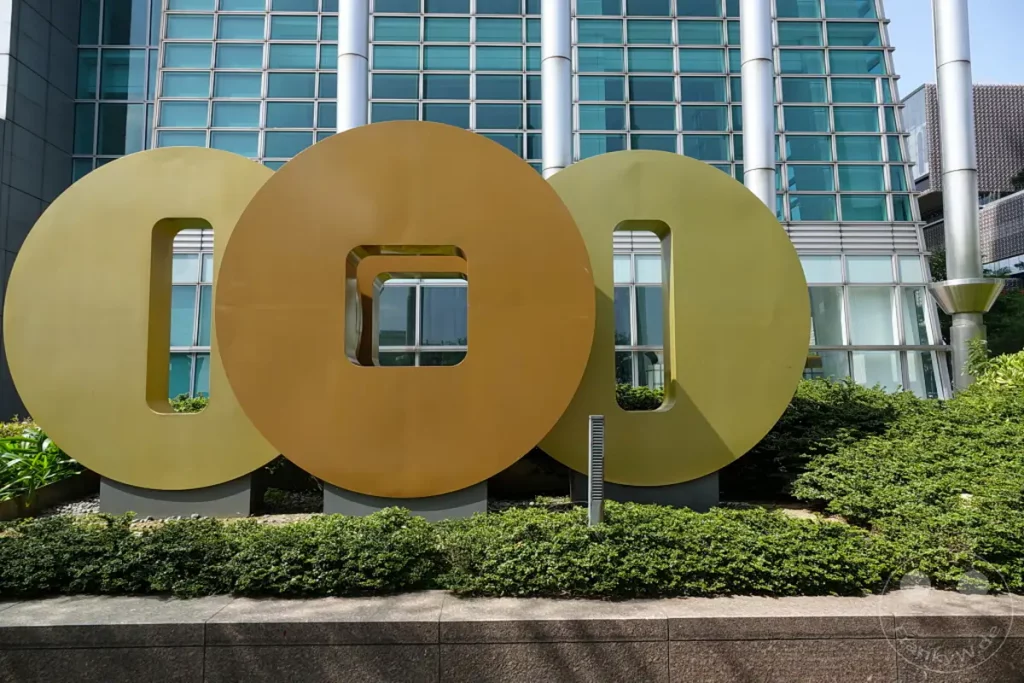 Vor der modernen Glasfassade des Taipei 101 in Taipeh, Taiwan, stehen drei riesige, kreisförmige Skulpturen, die an chinesische Münzen erinnern. Die gold- und bronzefarbenen Kunstwerke mit ihren rechteckigen und quadratischen Ausschnitten spiegeln die umliegende urbane Landschaft wider. Ein gepflegter grüner Buschstreifen bildet die Basis für die beeindruckende Installation.