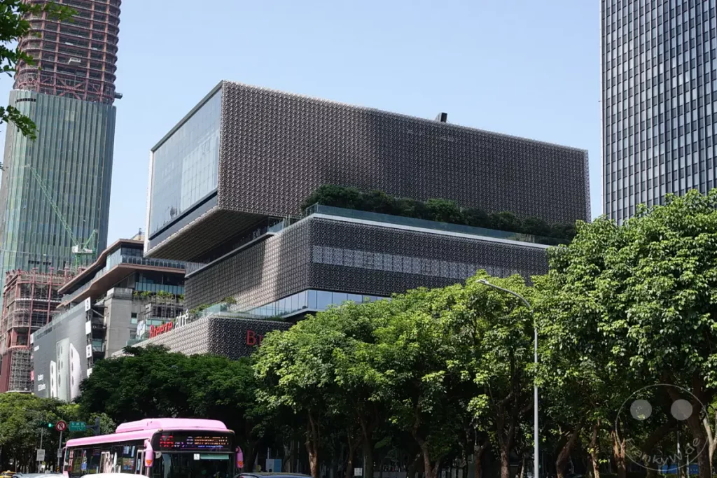 In Taipeh, Taiwan, ragt die beeindruckende Architektur des Taipei 101 im Hintergrund auf, während ein modernes Einkaufszentrum mit seiner markanten Fassade im Vordergrund dominiert. Eine leuchtend pinke Buslinie durchquert die breite Straße, gesäumt von üppigem Grün, das der urbanen Szene eine lebendige Atmosphäre verleiht. Die Szene vermittelt einen Eindruck von einer pulsierenden Metropole, in der moderne Baukunst und städtisches Leben harmonisch koexistieren.