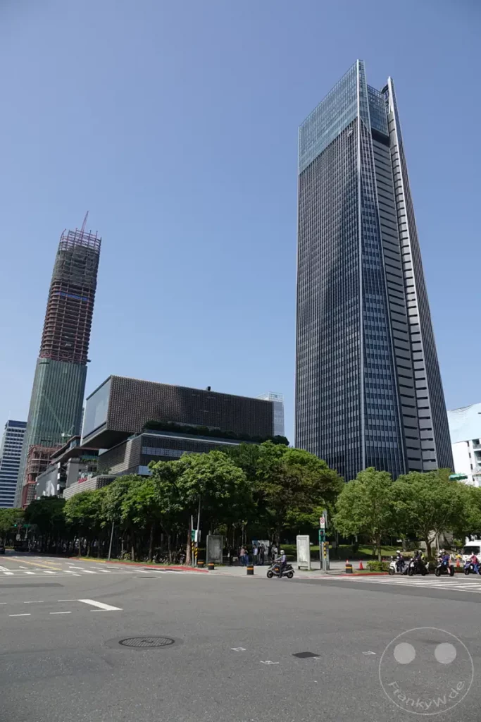 In Taipeh, Taiwan, ragen moderne Wolkenkratzer wie der markante Taipei 101 in den strahlend blauen Himmel. Unterhalb der imposanten Gebäude erstrecken sich grüne Bäume und eine belebte Straßenkreuzung, auf der Rollerfahrer unterwegs sind. Die Architektur der Stadt spiegelt eine dynamische Mischung aus fortschrittlichem Design und urbanem Leben wider.