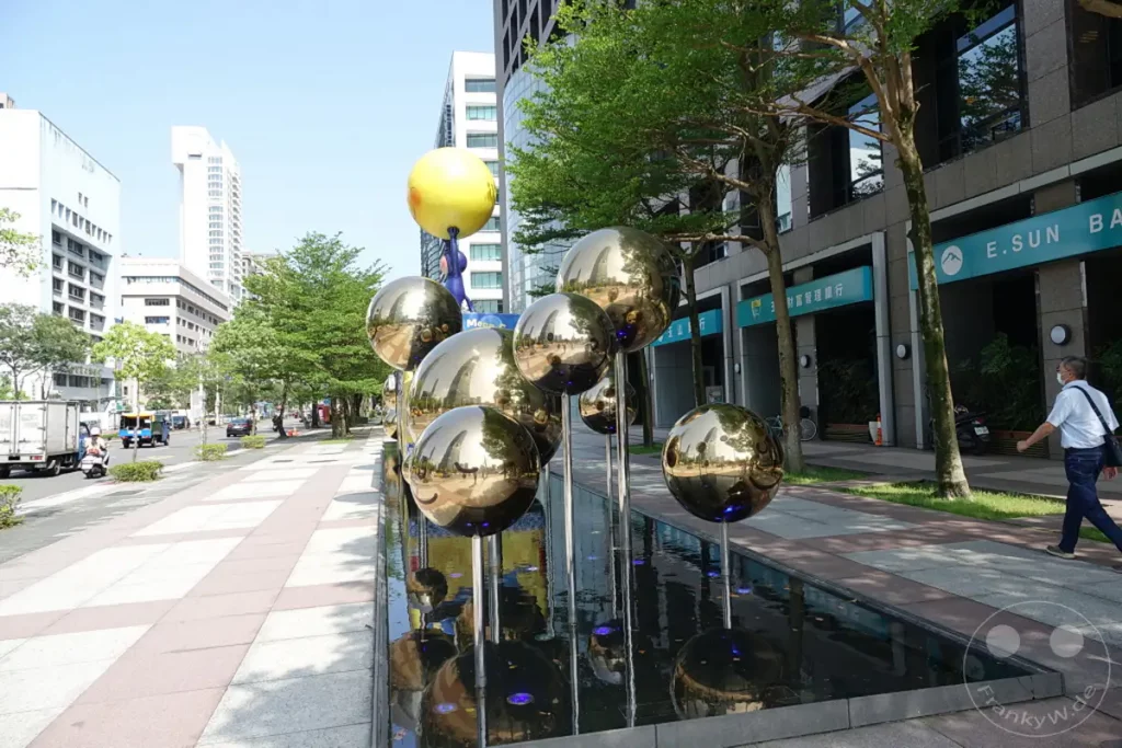 In Taipeh, Taiwan, steht eine moderne Kunstinstallation mit dem Titel "When the Moon forgot", die mehrere glänzende Kugeln in einem Wasserbecken zeigt. Im Hintergrund erheben sich moderne Gebäude, darunter der imposante Taipei 101, und ein belebter Straßenverkehr sorgt für urbanes Flair. Die Szene ist von strahlendem Sonnenschein geprägt und vermittelt eine lebendige Atmosphäre der Metropole.