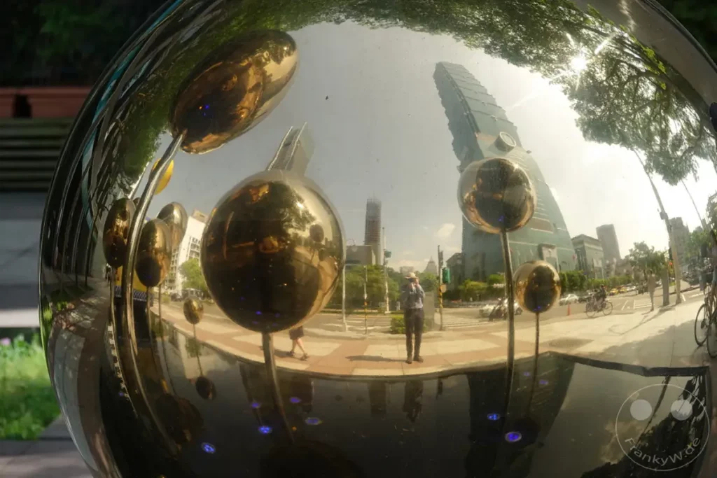 Eine polierte kugelförmige Skulptur reflektiert die städtische Umgebung von Taipeh, Taiwan, einschließlich des ikonischen Taipei 101 Wolkenkratzers und der umliegenden Architektur. Die Reflexionen sind verzerrt und spiegeln die Dynamik und das Aussehen der Stadt auf künstlerische Weise wider. Die Skulptur "When the Moon forgot" fügt eine surreale und träumerische Note hinzu, die durch das Lichtspiel verstärkt wird.