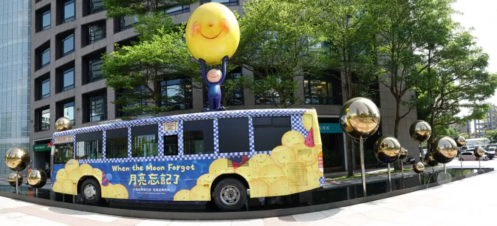 In Taipeh, Taiwan, steht ein künstlerisch gestalteter "Moon Bus" vor einem modernen Gebäude, das von reflektierenden Kugelskulpturen und grünen Bäumen gesäumt wird. Die Skulptur des Busses mit dem Titel "When the Moon Forgot" und eine Darstellung eines kleinen Wesens, das den Mond hält, erzeugen eine fantasievolle und verspielte Atmosphäre. Die gesamte Szenerie vor der Kulisse von städtischer Architektur und Natur strahlt eine fröhliche und neugierige Stimmung aus.