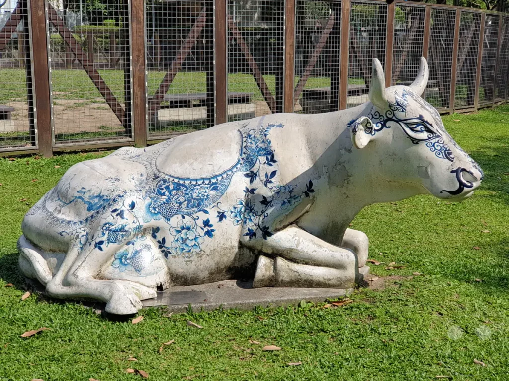 Eine weiße Kuh-Skulptur mit blauen Verzierungen liegt friedlich auf grünem Rasen in Taipeh, Taiwan. Im Hintergrund ist ein Zaun und ein Gebäude zu sehen, das an den Taipei 101 erinnert. Die Szene strahlt eine ruhige und künstlerische Atmosphäre aus.