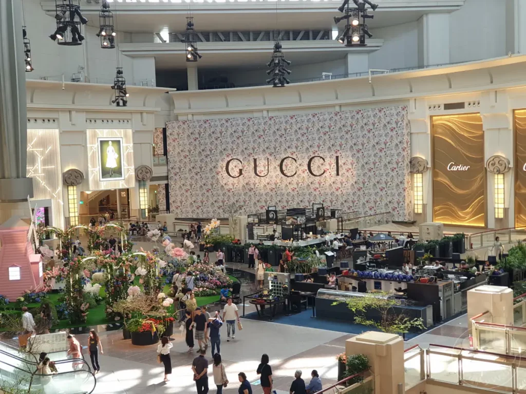 Ein Blick in das Innere eines luxuriösen Einkaufszentrums in Taipeh, Taiwan, mit dem markanten Gucci-Banner und der goldenen Cartier-Fassade. Eine üppige Blumeninstallation im Vordergrund schafft eine festliche Atmosphäre, während zahlreiche Menschen auf den verschiedenen Ebenen des Centers unterwegs sind.