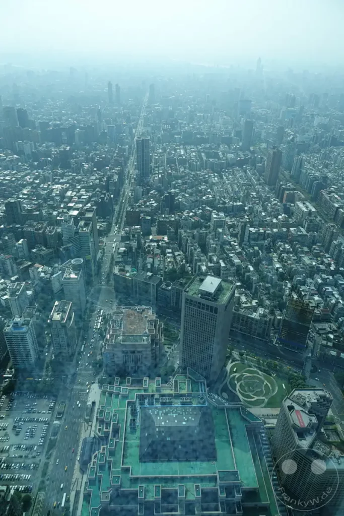 Eine weitläufige Stadtansicht von Taipeh, Taiwan, erfasst aus großer Höhe. Dichte Bebauung und moderne Wolkenkratzer prägen das Bild, während eine leichte Dunstschicht die ferne Skyline weichzeichnet. Die Aufnahme vermittelt ein Gefühl der urbanen Weite und des geschäftigen Treibens der Metropole.