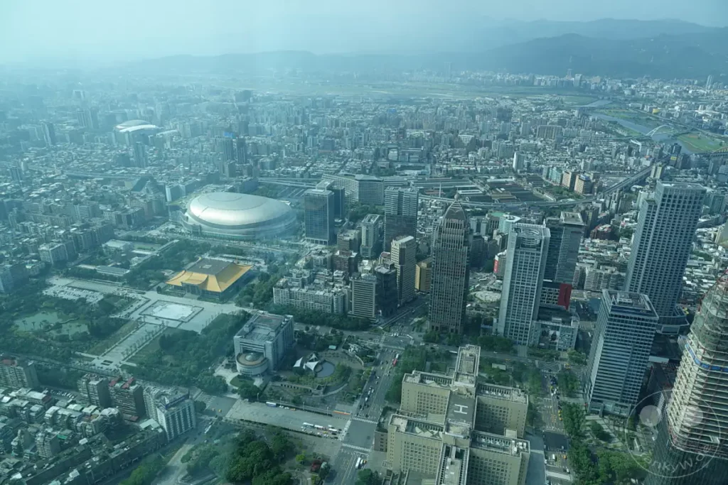 Eine weite Luftaufnahme erfasst die dichte Bebauung von Taipeh, Taiwan, mit zahlreichen Hochhäusern und weitläufigen Straßen. Im Vordergrund hebt sich ein großer Gebäudekomplex mit goldenem Dach ab, während im Hintergrund die markante Silhouette des Taipei 101-Turms die Skyline dominiert. Die Szene vermittelt einen Eindruck von der urbanen Größe und Geschäftigkeit der Metropole, eingefangen an einem leicht diesigen Tag.