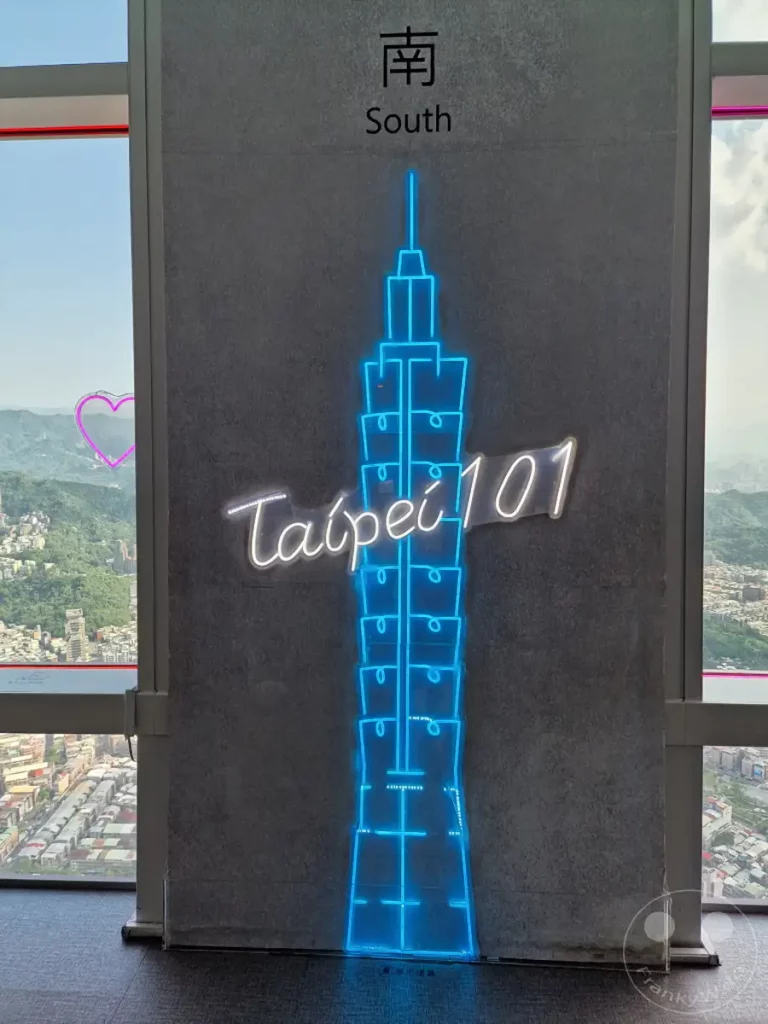 Die Neonlicht-Silhouette des Taipei 101 Gebäudes leuchtet eindrucksvoll an einer Wand in Taipeh, Taiwan. Draußen bietet sich ein weiter Blick über die Stadt und die umliegenden grünen Hügel. Eine herzförmige Lichterkette im Fenster ergänzt die stimmungsvolle Atmosphäre dieses Aussichtspunktes.