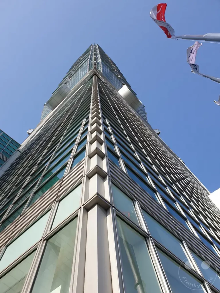 Die steil aufragende Fassade des Taipei 101 Wolkenkratzers dominiert den klaren blauen Himmel über Taipeh, Taiwan. Glas und Stahl spiegeln das Sonnenlicht wider und erzeugen ein Gefühl von Höhe und Erhabenheit. Drei wehende Flaggen am oberen Bildrand verleihen der städtischen Landschaft einen Hauch von Farbe und Bewegung.